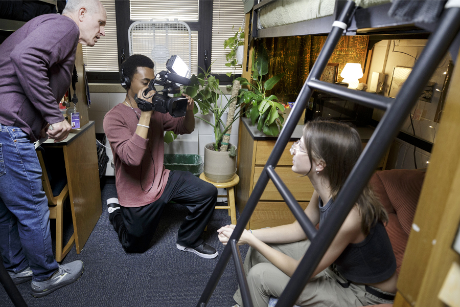 A student interviews another student in a dorm room with a video camera as a faculty member assists.